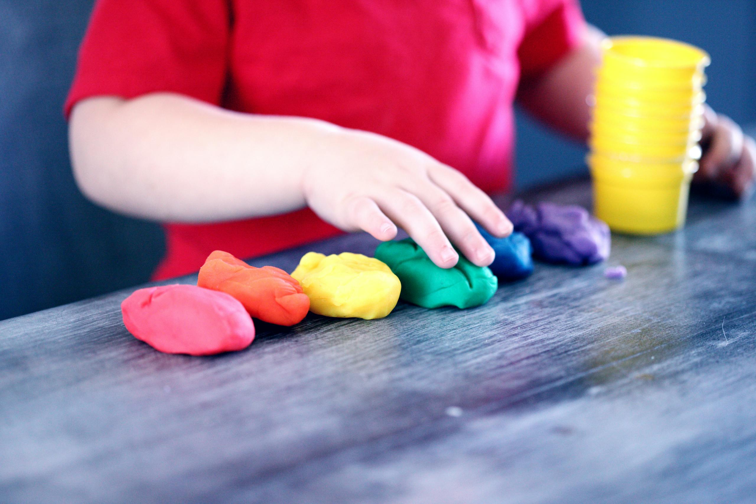 Hands of young child resting on a table with colours (pink, red, yellow green, blue and purple) of playdoh in front of them alongside a yellow cup.