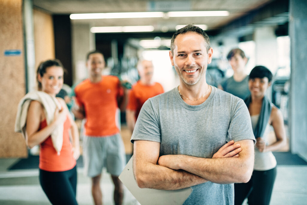 Man with clipboard in gym smiling at camera. Gym goers in background.
