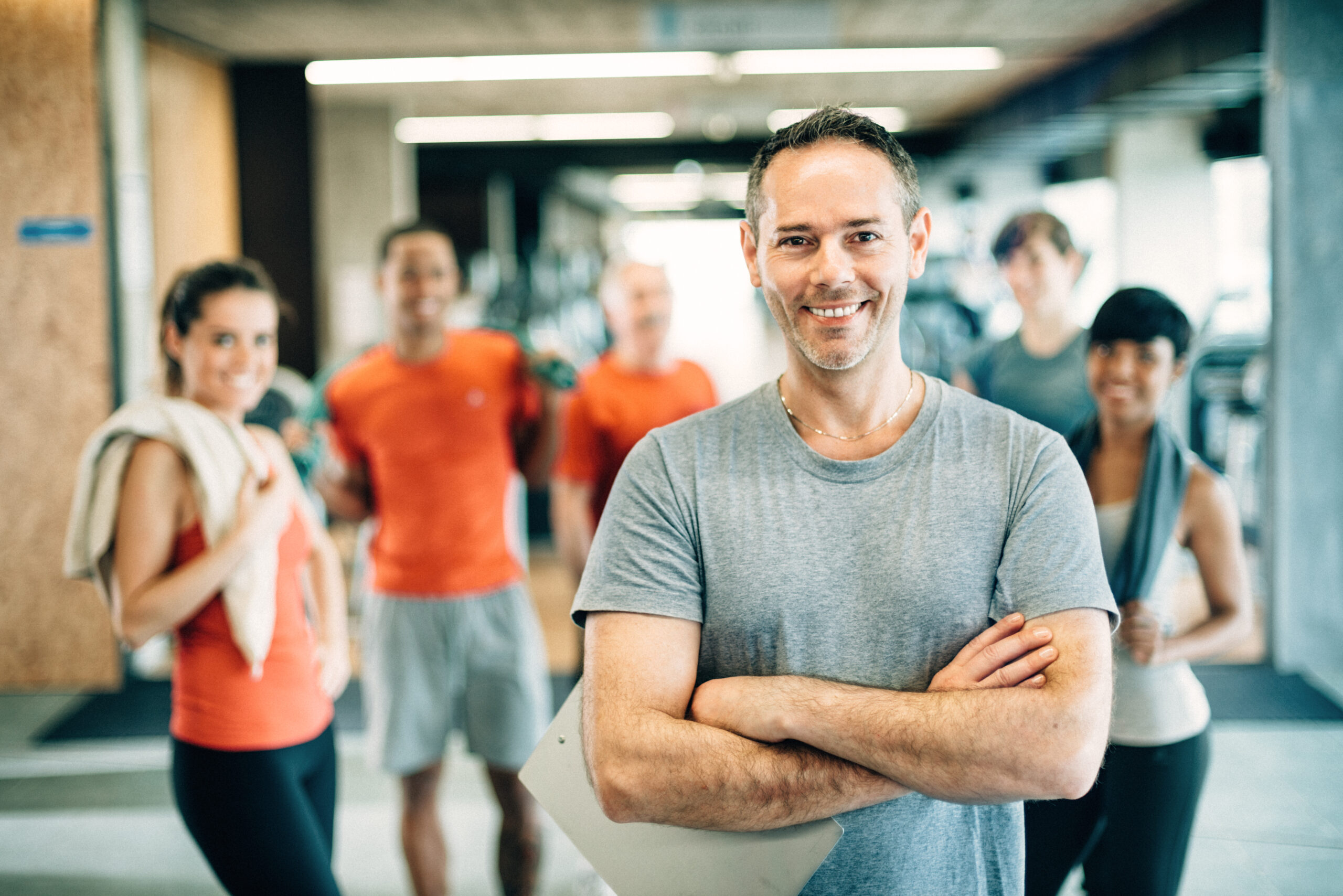 Man with clipboard in gym smiling at camera. Gym goers in background.